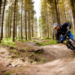 A man on a mountain bike cycles round a banked curve on a dirt track in a wooded area.