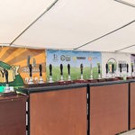 A long row of hand-pull beer taps arranged on wooden counters beneath a white marquee canopy. Colourful banners advertising different breweries and beers hang behind the taps.