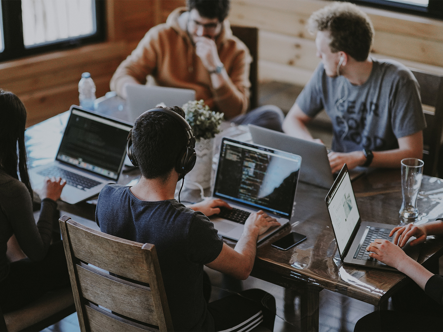 Five remote workers in casual dress sit round a large table, working on laptop computers.