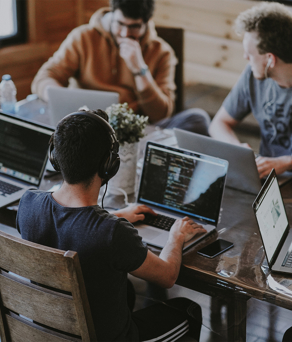 Five remote workers in casual dress sit round a large table, working on laptop computers.