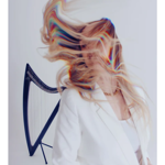 Person standing beside a harp against a plain light background, wearing a white blazer and light top. Long hair is swept dramatically across the face and upper body, creating a blurred, flowing effect with rainbow-like colour fringes. The harp is partially visible on the left, while the minimalist composition and bright lighting emphasise movement and the artistic, contemporary style of the portrait.