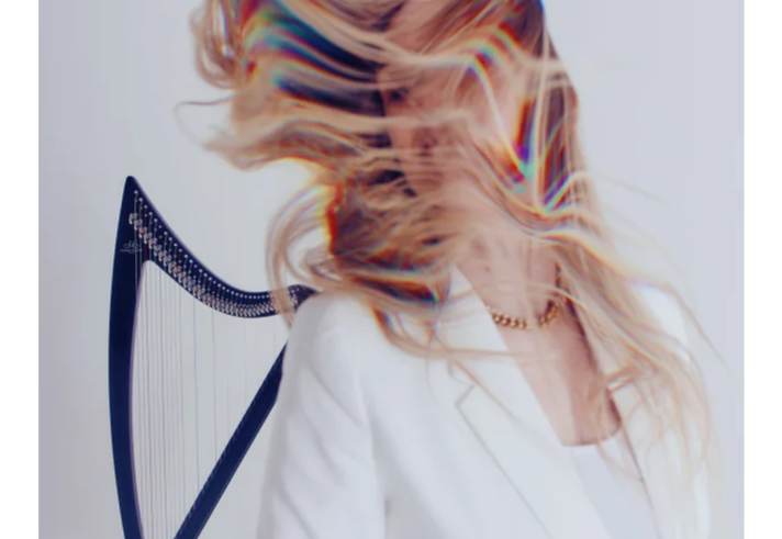 Person standing beside a harp against a plain light background, wearing a white blazer and light top. Long hair is swept dramatically across the face and upper body, creating a blurred, flowing effect with rainbow-like colour fringes. The harp is partially visible on the left, while the minimalist composition and bright lighting emphasise movement and the artistic, contemporary style of the portrait.