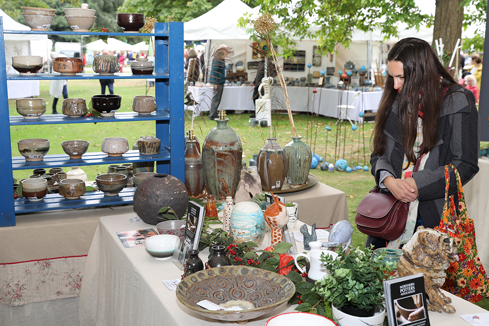 A pottery stall at an outdoor craft fair displays a variety of handmade ceramic items. A blue wooden shelf holds rows of bowls in different sizes and earthy tones. The table in front is covered with beige fabric and features large decorative vases, a patterned dish, small pots, and green plants. Leaflets and business cards are scattered on the table. In the background, white tents and other stalls are visible under leafy trees.