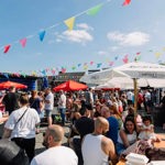  A lively outdoor event at a pub beer garden, with crowds sitting at picnic tables under colourful bunting and large umbrellas on a bright sunny day.