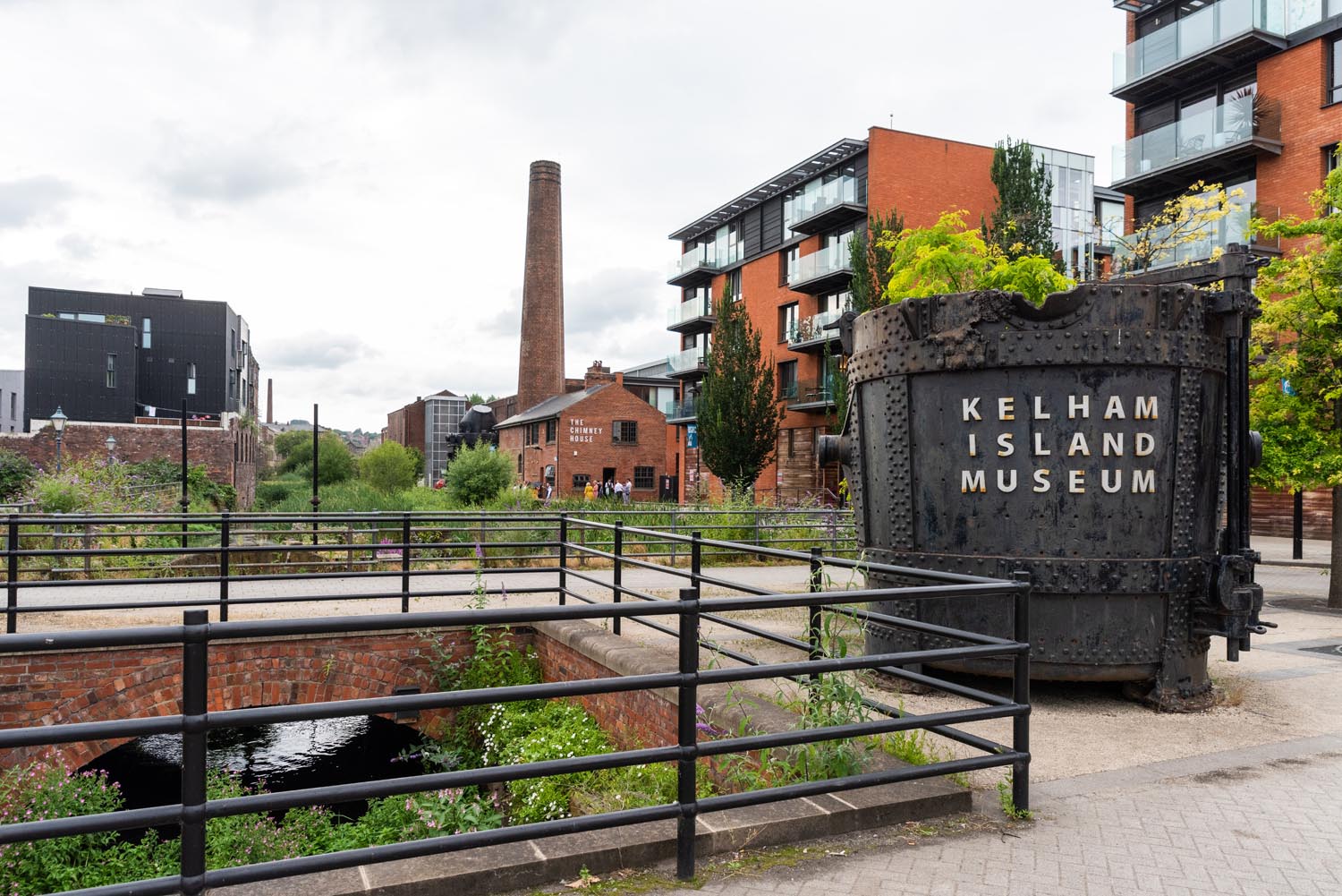 Outdoor view of Kelham Island Museum in Sheffield. A large industrial metal container with “Kelham Island Museum” written on it stands in the foreground near railings and a water channel. Surrounding the area are modern apartment buildings, a tall brick chimney, and greenery under a cloudy sky.