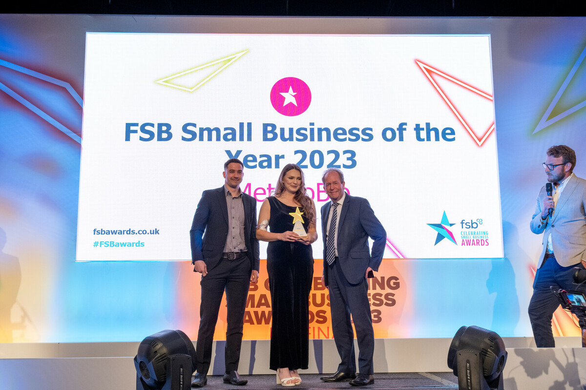 Three people standing on a stage at an awards ceremony with a large screen behind them displaying the text: “FSB Small Business of the Year 2023” and “fsbawards.co.uk #FSBAwards.” The person in the center is holding a trophy, while the two individuals on either side are dressed in formal attire. A fourth person with a microphone is positioned to the right, appearing to host or announce. The stage is brightly lit with colourful geometric designs in the background.