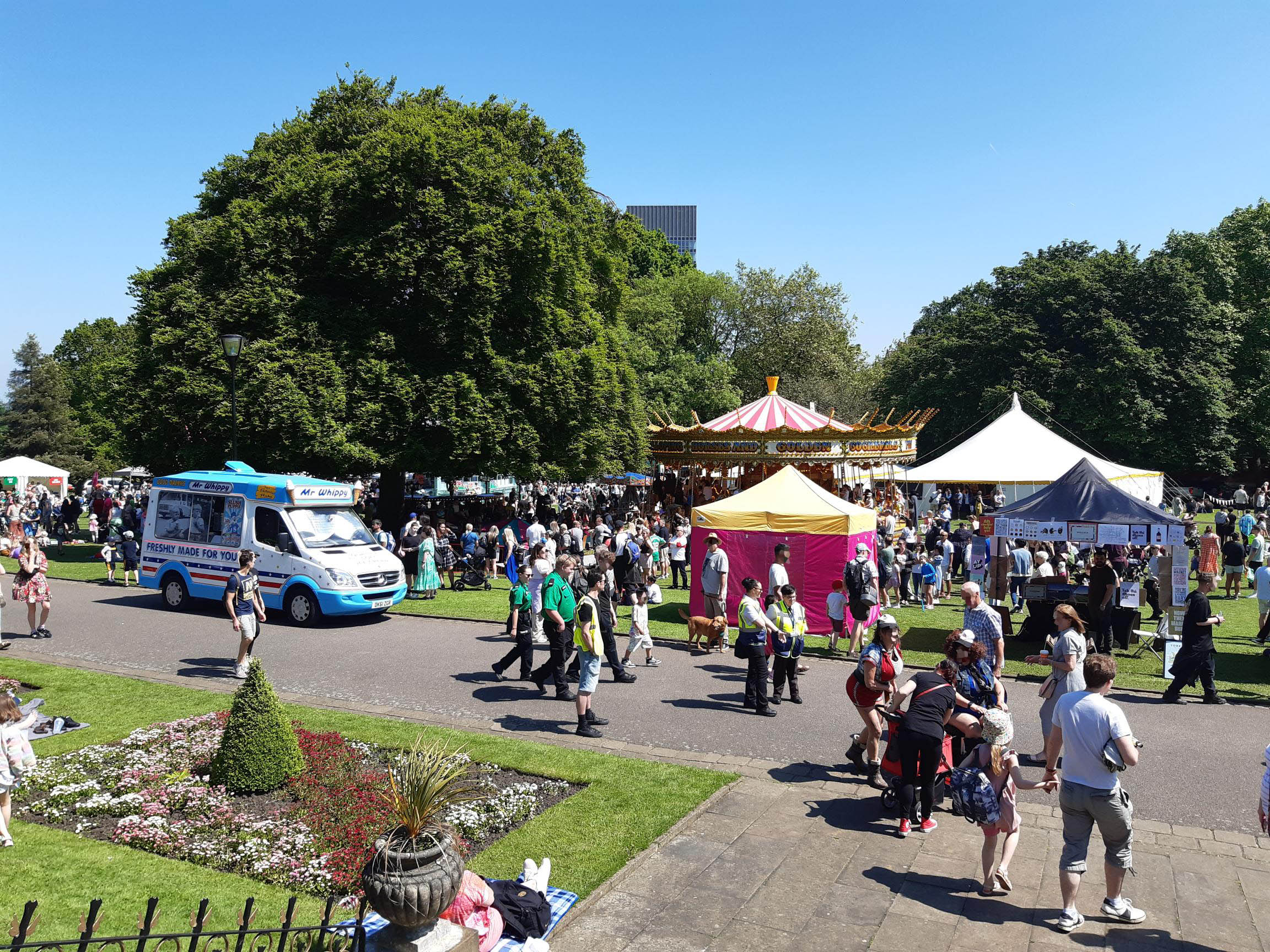 A busy outdoor festival scene in a park on a sunny day. Crowds of people walk and gather near colourful tents, including a bright yellow and pink stall, and a traditional carousel with a striped canopy. An ice cream van is parked on a paved path to the left. The foreground shows a flowerbed with neatly trimmed greenery, while large trees provide shade in the background.