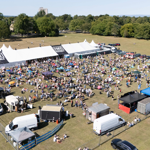 A large outdoor music festival set in an open grassy field with a white marquee tent labeled “FAKE FESTIVALS.” Crowds of people are gathered in front of the tent, sitting on blankets or standing near the stage area. Surrounding the festival are food stalls, trucks, and inflatable attractions, with trees and countryside visible in the background under a clear blue sky.