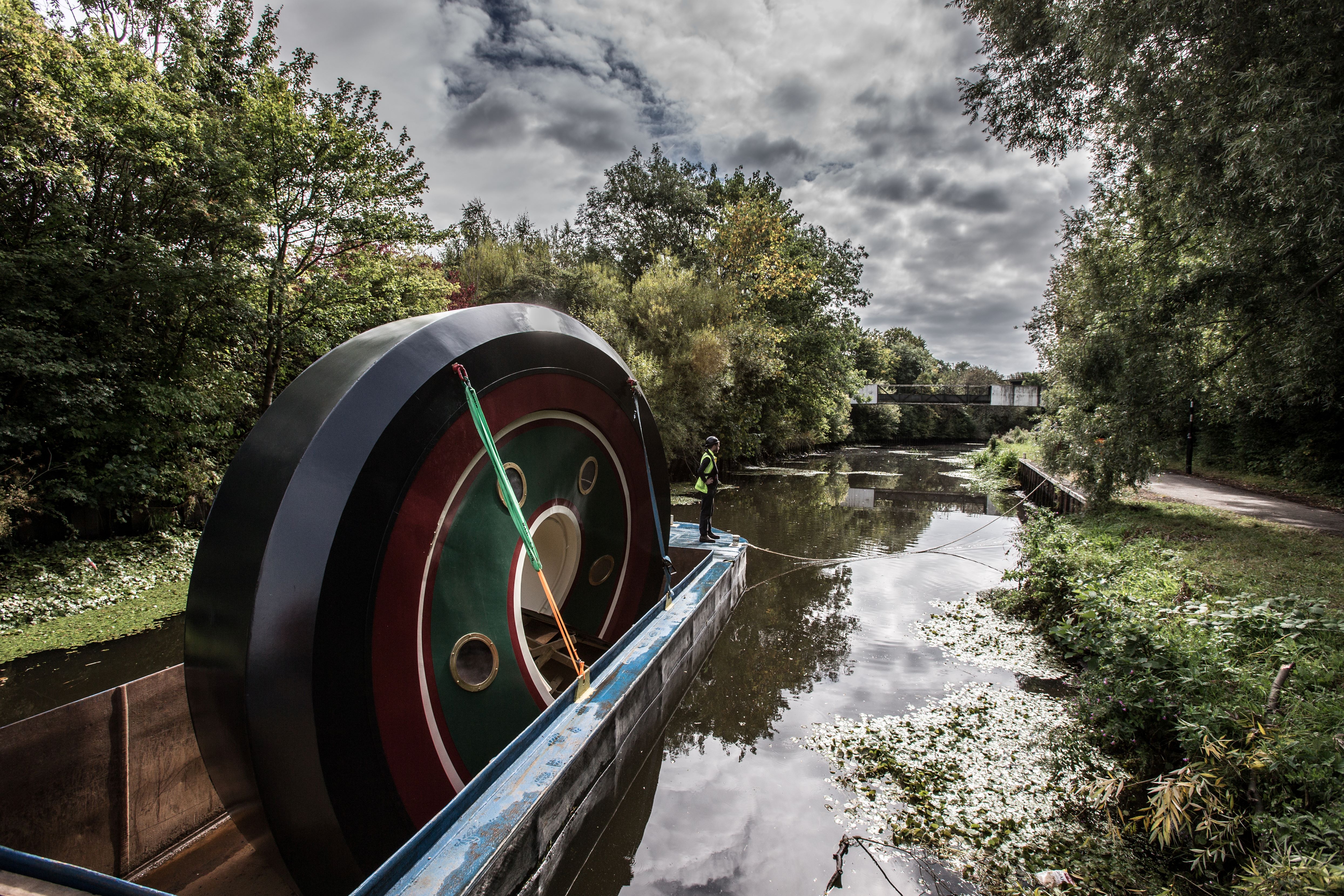 Large circular sculpture with green, red, and black rings being transported on a flat barge along a narrow canal, surrounded by dense trees and vegetation under a cloudy sky.