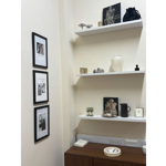 Corner display in a minimalist boutique or gallery space. Three black-framed black-and-white photographs hang on a cream wall beside white floating shelves holding decorative objects, books, glassware and ceramics. Visible titles include books on Henry Moore and Rodin. A dark wooden cabinet below displays stacked cards and a shallow dish. The neutral colour palette and carefully arranged objects create a refined, gallery-like atmosphere.