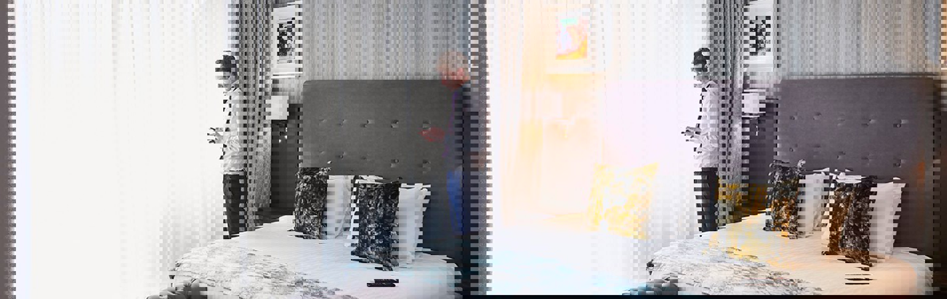 Elegant luxury bedroom featuring plush cushions on a king-sized bed, soft ambient lighting, and a large window with sheer curtains letting in natural light.