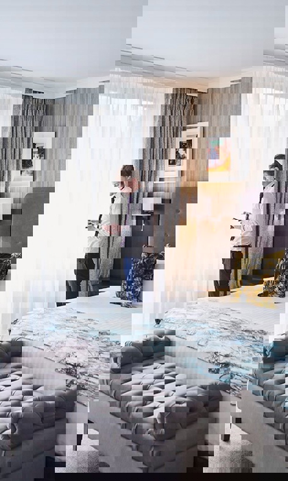 Elegant luxury bedroom featuring plush cushions on a king-sized bed, soft ambient lighting, and a large window with sheer curtains letting in natural light.