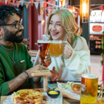 Two people are seated at a wooden table in a lively bar or beer hall, holding large glasses of beer and ale in a toast. The table has plates of food, including pretzels with mustard dip and other snacks. A red menu with the word “KAPITAL” is visible, and the background features colourful bunting, warm lighting, and a modern interior design.