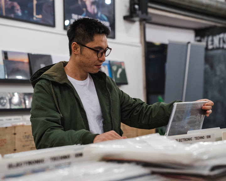 A person wearing a white T-shirt and a green jacket is browsing through vinyl records in a record store. The individual is holding one record while looking through a bin labeled with music genres such as "Electronic Techno" and "Psychedelic Rock." The background shows shelves with more records and framed artwork on the wall.