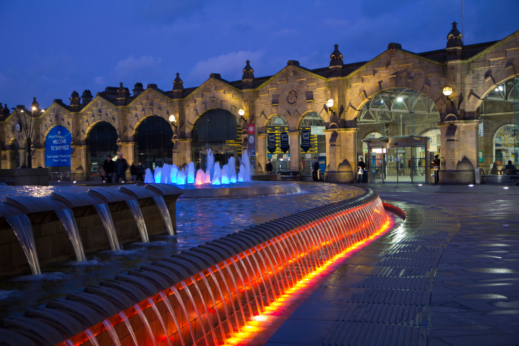 Illuminated water feature with cascading streams and glowing red lights in the foreground, set in front of a historic stone railway station building at dusk. The station has large arched windows and ornate architectural details, with colourful fountains lit in blue and purple near the entrance.