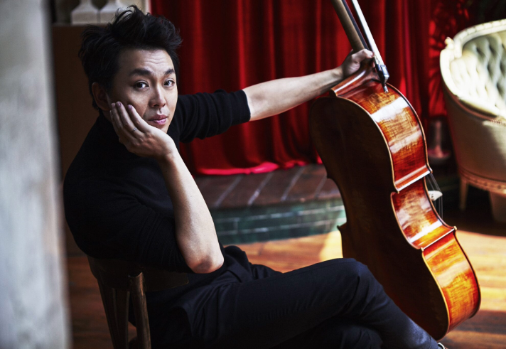A person dressed in black is seated on a wooden chair indoors, holding a cello upright by its neck. The cello has a polished wooden finish and rests on the floor beside the chair. The background features a red curtain and a light-coloured upholstered chair, with warm wooden flooring adding to the intimate setting.