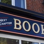 Close-up view of the Next Chapter Books shop sign, featuring bold cream lettering on a dark blue background with red and white borders. The phrases ‘WE BUY BOOKS’ and ‘NEXT CHAPTER BOOKS’ are displayed on the corner fascia of a red‑brick building.