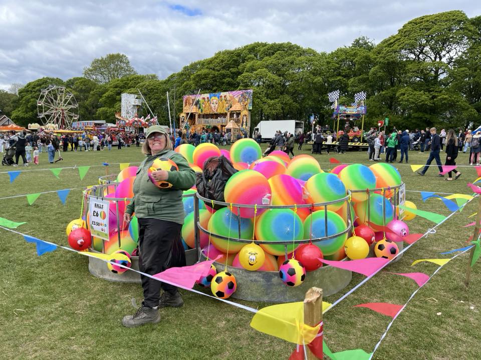  A colourful fairground stall featuring a circular pen filled with large rainbow-striped inflatable balls and smaller patterned balls. Bright bunting in pink, blue, green, and yellow surrounds the area. A sign on the pen reads “Balls £6 or 2 for £10.” In the background, crowds of people walk across the grassy field, with fairground rides, stalls, and trees visible under a partly cloudy sky.