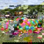  A colourful fairground stall featuring a circular pen filled with large rainbow-striped inflatable balls and smaller patterned balls. Bright bunting in pink, blue, green, and yellow surrounds the area. A sign on the pen reads “Balls £6 or 2 for £10.” In the background, crowds of people walk across the grassy field, with fairground rides, stalls, and trees visible under a partly cloudy sky.