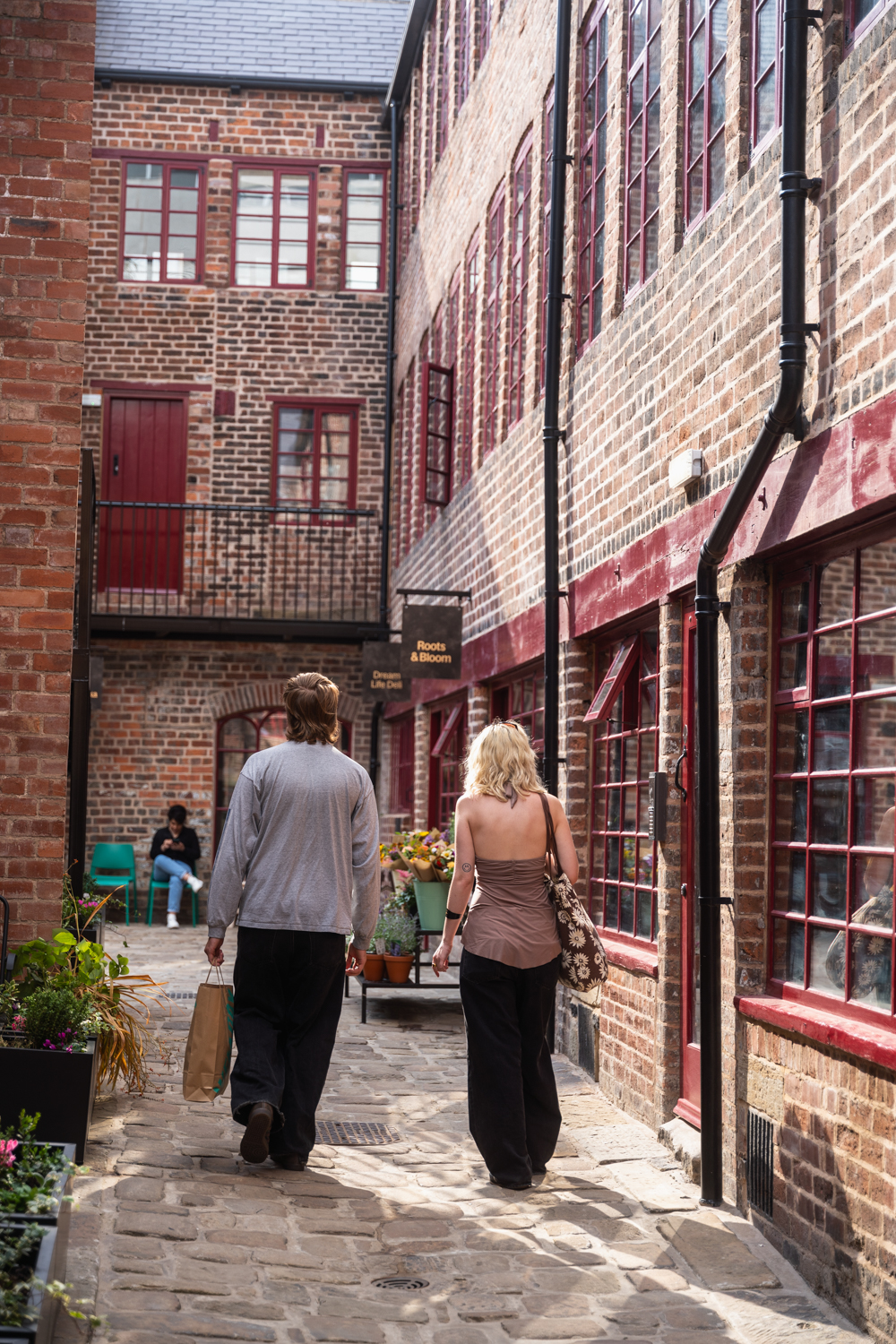 Two people walking along a cobbled courtyard lined with red‑brick buildings and small shopfronts, with potted plants, windows and doors creating a sheltered, historic city‑centre passageway.