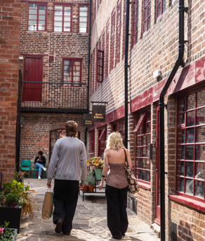 Two people walking along a cobbled courtyard lined with red‑brick buildings and small shopfronts, with potted plants, windows and doors creating a sheltered, historic city‑centre passageway.