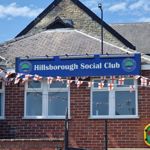 Brick building with a blue sign reading “Hillsborough Social Club” mounted above a row of white-framed windows. Red brick walls and a slate roof sit beneath a partly cloudy blue sky. Strings of small red-and-white flags are draped across the front, suggesting a celebration or event. A black pole stands near the sign, and a yellow defibrillator box is fixed to the right side of the wall. Steps with a metal handrail lead up toward the entrance, though the door is not visible.
