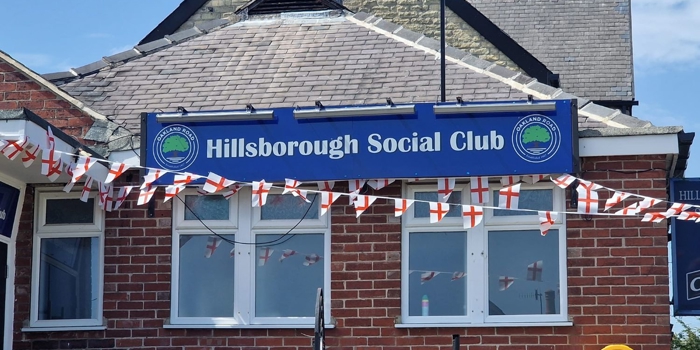 Brick building with a blue sign reading “Hillsborough Social Club” mounted above a row of white-framed windows. Red brick walls and a slate roof sit beneath a partly cloudy blue sky. Strings of small red-and-white flags are draped across the front, suggesting a celebration or event. A black pole stands near the sign, and a yellow defibrillator box is fixed to the right side of the wall. Steps with a metal handrail lead up toward the entrance, though the door is not visible.