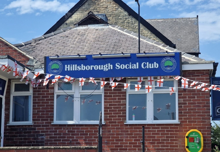 Brick building with a blue sign reading “Hillsborough Social Club” mounted above a row of white-framed windows. Red brick walls and a slate roof sit beneath a partly cloudy blue sky. Strings of small red-and-white flags are draped across the front, suggesting a celebration or event. A black pole stands near the sign, and a yellow defibrillator box is fixed to the right side of the wall. Steps with a metal handrail lead up toward the entrance, though the door is not visible.