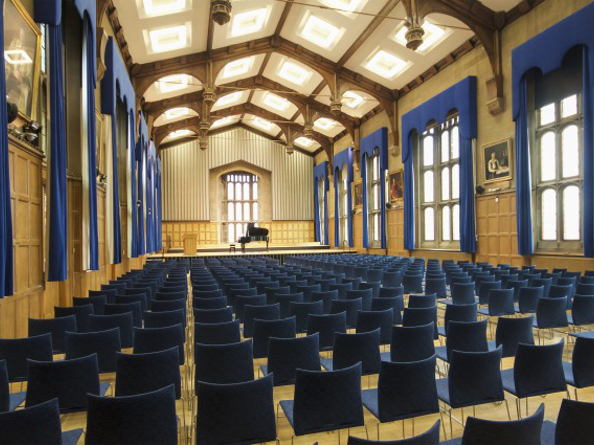 Interior of a grand hall with high wooden-beamed ceiling and large arched windows draped in blue curtains. Rows of blue chairs face a raised stage with a black grand piano, and the walls feature wood panelling and framed portraits.