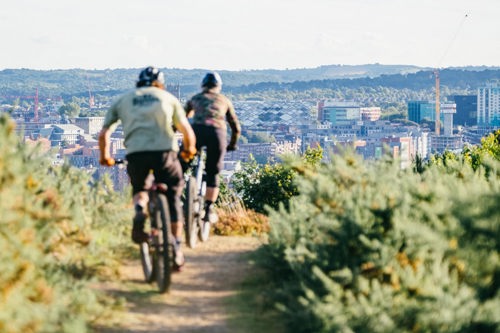 Two cyclists riding mountain bikes along a dirt trail surrounded by green shrubs. The path leads toward a scenic view of a city in the background, with buildings, cranes, and rolling hills visible under a clear sky. The image captures an outdoor adventure with an urban landscape in the distance.