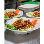 Three plated servings of a glossy brown stir-fried dish are arranged on a stainless-steel kitchen counter. The foreground plate is garnished with fresh coriander, shredded cucumber, carrot and sesame seeds, while two similar plates sit slightly out of focus in the background. The image highlights the presentation and preparation of the meal in a restaurant kitchen.