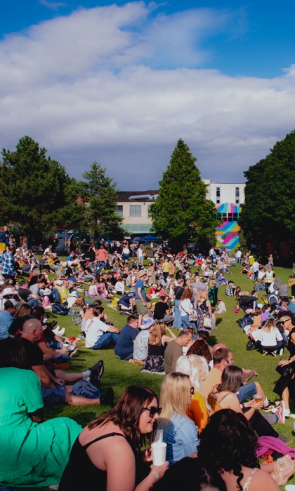 A big crowd of people are sitting on an open are of grass in the sunshine, enjoying an outdoor event in the centre of Sheffield.