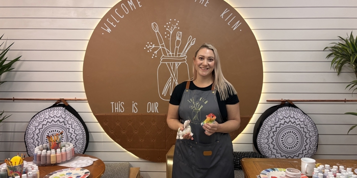 Person wearing a gray apron standing in front of a circular sign that reads ‘Welcome to the Kiln’ and ‘This is our…’ with an illustration of paintbrushes. The setting includes wooden tables with paint bottles, ceramic items, and color palettes arranged for a pottery painting activity. Decorative cushions and potted plants are visible in the background.