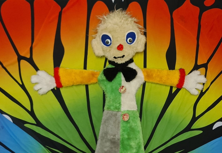 A colourful hand puppet with fluffy cream hair, large blue eyes and a red nose is displayed with its arms stretched wide. The puppet wears a patchwork outfit in green, white, grey and yellow.