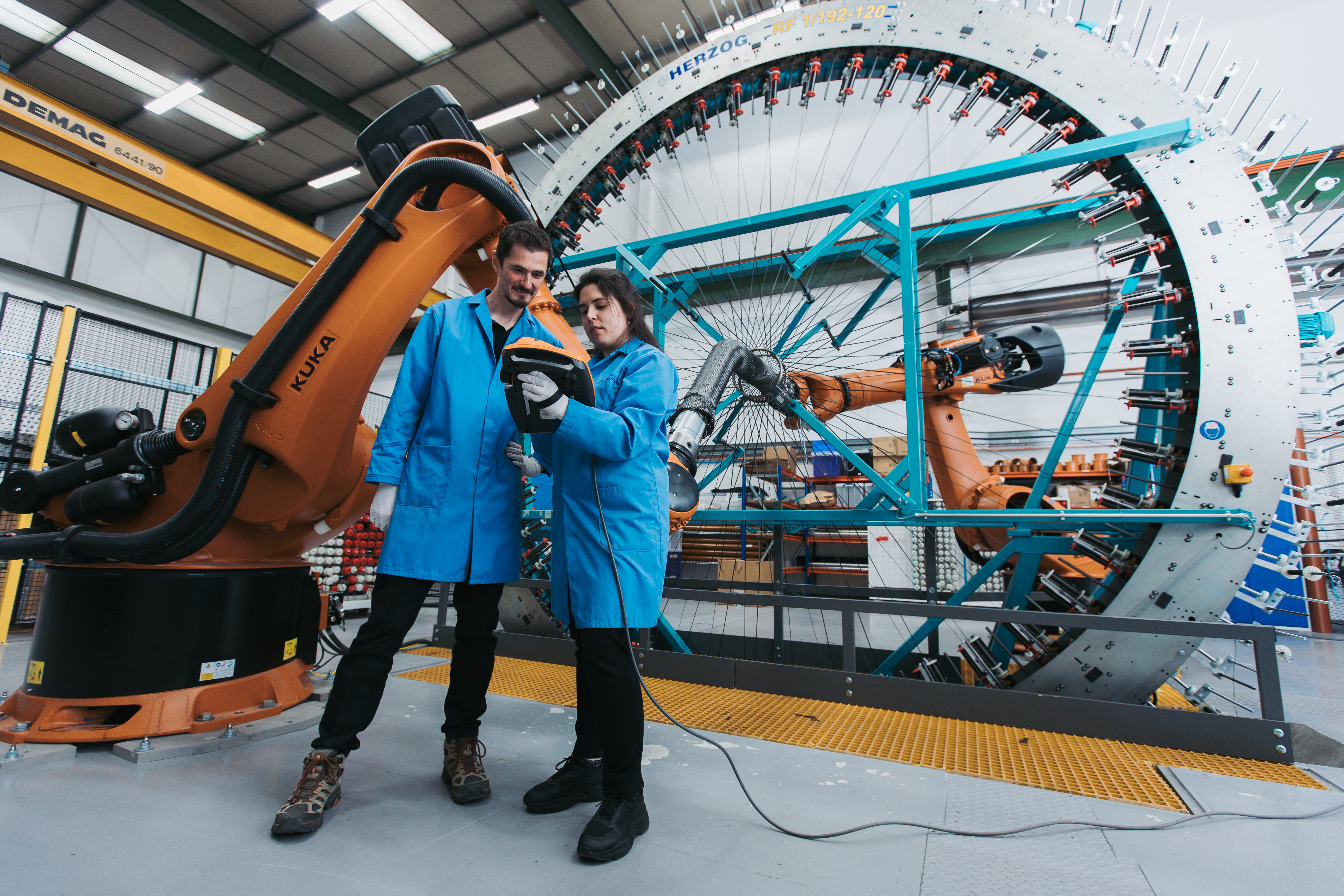 Two people in blue lab coats are talking and looking at a tablet like device. Behind them is an industrial weaving loom.