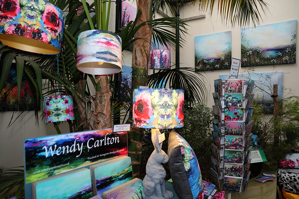 A vibrant indoor art display featuring colourful abstract paintings on the walls, patterned lampshades hanging and on stands, and matching cushions. A sign reads “Wendy Carlton” with promotional material below. A rack holds greeting cards with similar artwork, and decorative items, including two sculpted rabbits, are arranged on a table. The setting includes lush green plants, creating a lively and artistic atmosphere.