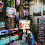 A vibrant indoor art display featuring colourful abstract paintings on the walls, patterned lampshades hanging and on stands, and matching cushions. A sign reads “Wendy Carlton” with promotional material below. A rack holds greeting cards with similar artwork, and decorative items, including two sculpted rabbits, are arranged on a table. The setting includes lush green plants, creating a lively and artistic atmosphere.