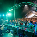 A live band performs under a large tent with bright green stage lights illuminating the musicians and equipment. The guitarist stands near the front, pointing toward a packed audience behind a barrier. The crowd is cheering with raised hands, and the stage setup includes speakers, fans, and lighting rigs overhead.