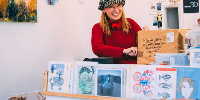Inside Cupola Gallery at Hillsborough. The walls are covered with paintings by local artists. In the foreground is a display stand filled with prints and greetings cards. A smiling woman is standing in the background opening a large package.