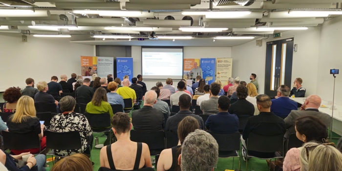 A large group of people seated in rows facing a presentation screen in a modern conference room. Several banners are displayed at the front near the screen, featuring business and enterprise branding. The presenter stands near the screen, which shows a slide with text. The room has bright lighting, white walls, and exposed ceiling fixtures.