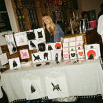 Indoor market stall displaying cat-themed artwork and merchandise. A person stands behind a table covered with prints, cards and tote bags featuring black cat illustrations with pink and red accents. Wire display racks hold framed and mounted artworks, while a bar area with drinks shelves, menu boards and string lights forms the background. A clothing rack with white T-shirts stands to one side.