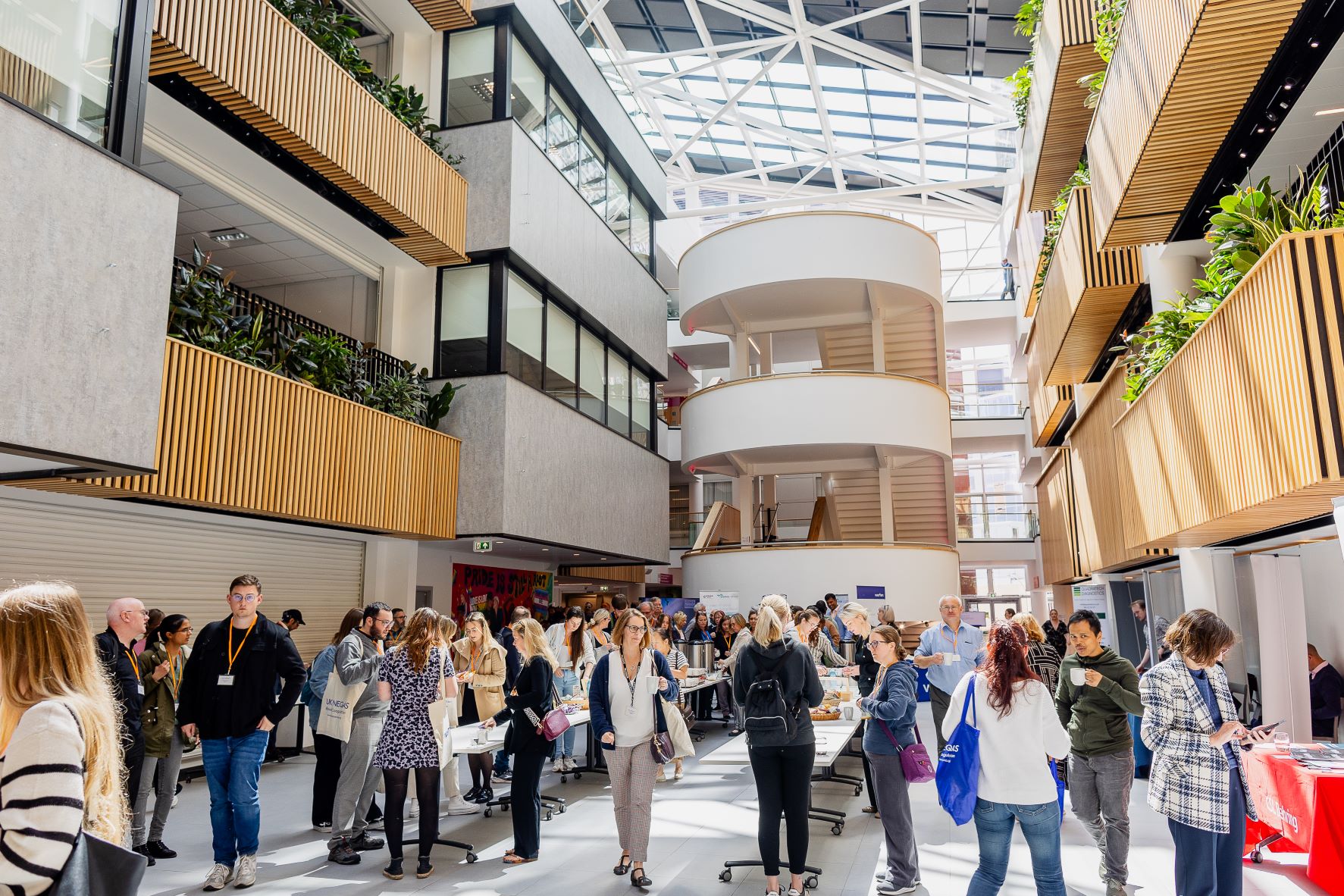 A bright, modern atrium with high glass ceilings and wooden panel accents on the walls. The space is filled with people standing and walking around tables, suggesting a networking event or conference. Several floors with balconies and greenery overlook the central area, and natural light streams in from the large skylight above.