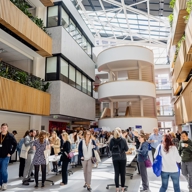 A bright, modern atrium with high glass ceilings and wooden panel accents on the walls. The space is filled with people standing and walking around tables, suggesting a networking event or conference. Several floors with balconies and greenery overlook the central area, and natural light streams in from the large skylight above.