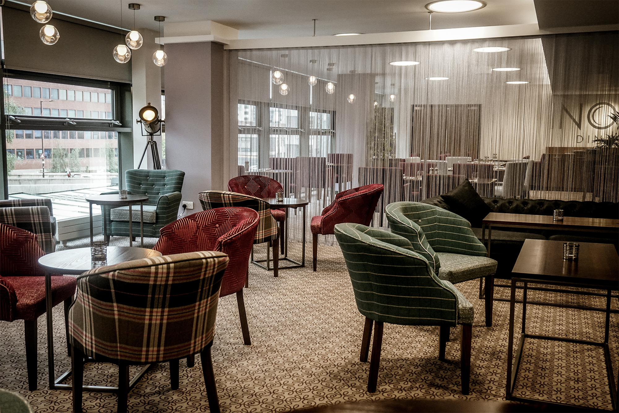 The interior of Inox Dine. Modern lounge area with patterned carpet, round tables, and upholstered chairs in various plaid and striped designs. The space features large windows providing natural light, a decorative floor lamp, and a semi-transparent curtain dividing the room. In the background, there are more tables and chairs arranged for dining, with pendant lights hanging from the ceiling.