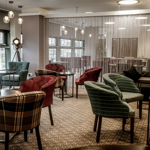 The interior of Inox Dine. Modern lounge area with patterned carpet, round tables, and upholstered chairs in various plaid and striped designs. The space features large windows providing natural light, a decorative floor lamp, and a semi-transparent curtain dividing the room. In the background, there are more tables and chairs arranged for dining, with pendant lights hanging from the ceiling.