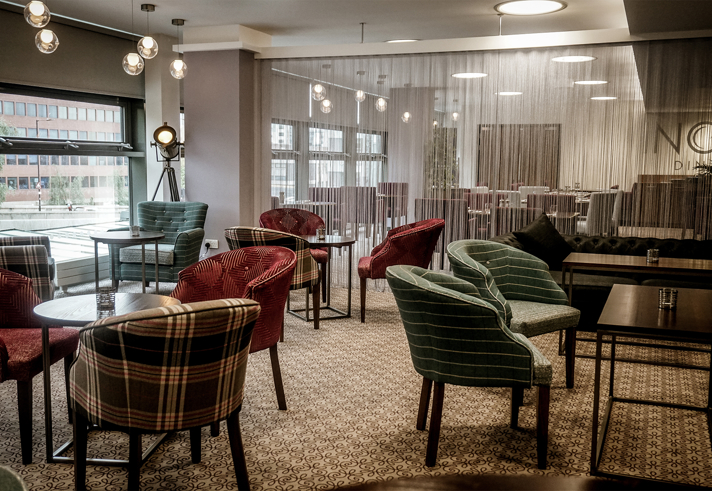 The interior of Inox Dine. Modern lounge area with patterned carpet, round tables, and upholstered chairs in various plaid and striped designs. The space features large windows providing natural light, a decorative floor lamp, and a semi-transparent curtain dividing the room. In the background, there are more tables and chairs arranged for dining, with pendant lights hanging from the ceiling.