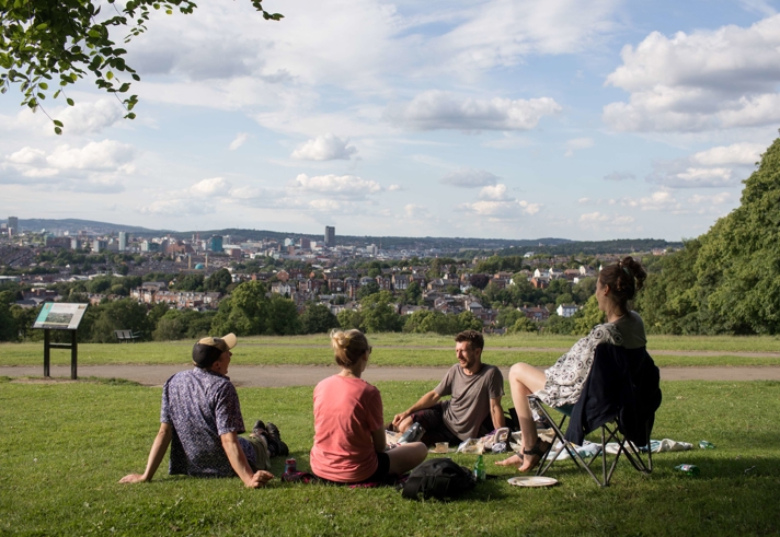 Four people are sat on the grass at Meersbrook Park in the sun. In the distance you can see Sheffield city centre.