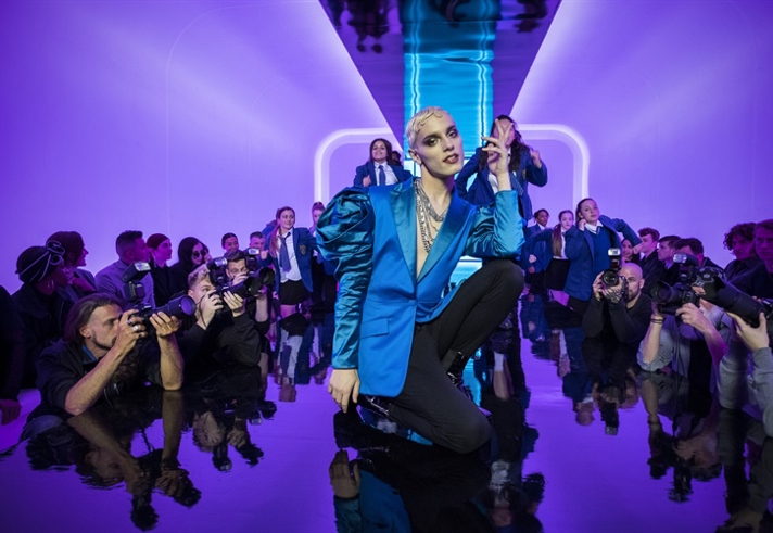 A person in a bold blue outfit with exaggerated shoulders poses confidently on a reflective runway. Purple lighting illuminates the background, while photographers surround the runway, capturing the moment at what appears to be a fashion show.