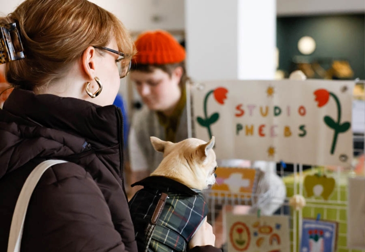 A person wearing glasses and a dark jacket holds a small dog in a plaid coat in front of a booth labeled "STUDIO PHEEBS," decorated with colorful flowers. Another person in an orange beanie stands behind the booth, which displays various art or craft items, suggesting a lively market or fair setting.
