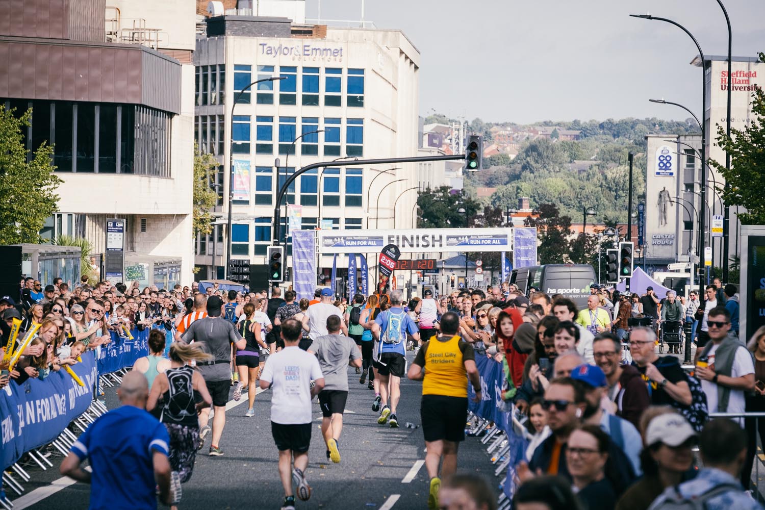 A group of people running towards the finish line of Sheffield Half Marathon in Castlegate on a sunny day. Spectators are cheering on from either side of the track.